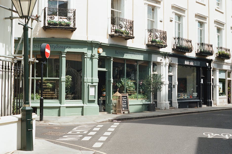 A storefront of a florist shop with a pastel green facade, displaying fresh flowers in the window. The shop has large glass windows, and potted plants are visible inside. Outside, there is a black cha