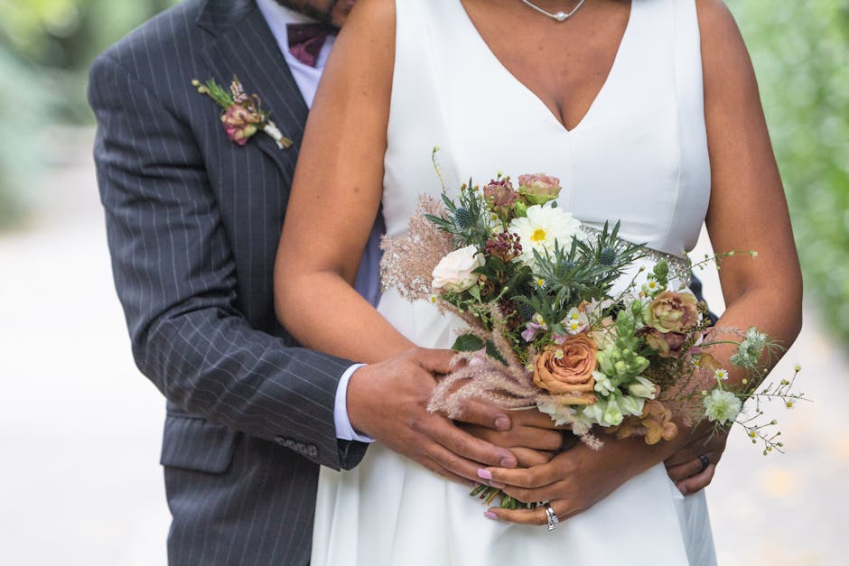 Close-up of a bride and groom holding a floral bouquet with soft peach, white, and mauve roses, interspersed with greenery and delicate filler flowers, wrapped in pastel pink tissue paper. The bride's