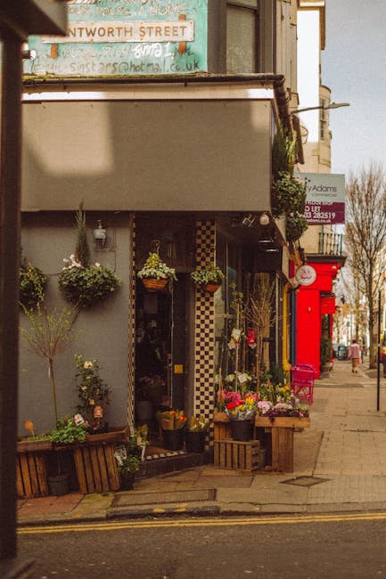 A charming floral display outside a flower shop on a street corner, featuring vibrant, fresh flowers arranged in wooden crates and hanging baskets. The bouquet includes bright pink roses, yellow and w