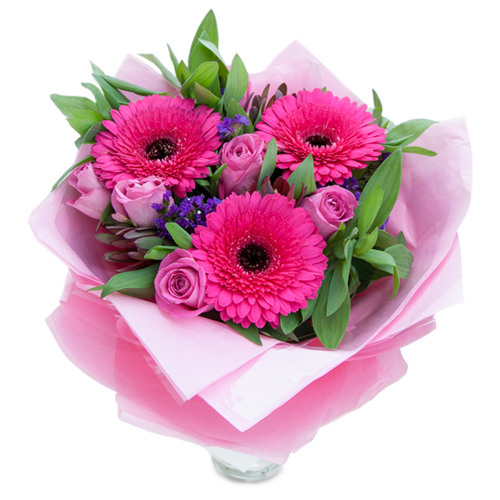 I picture the composition from a florist's perspective: three confident Gerbera daisies in a striking fuchsia sit proudly at the centre, their layered petals fanning outward like small, sunlit rosettes, each with a dark, defining centre that gives the arrangement a note of contrast and structure. Tucked in close are several roses in a muted lavender-pink, petals still gently cupped as if protecting a tender scent-soft, powdery and floral. Sprigs of purple statice thread through the bouquet, introducing a papery, textural element and a cool violet whisper that balances the warmer pinks. The greenery-broad, glossy leaves that recall salal with hints of aromatic eucalyptus-provides a deep, verdant canvas, its fresh, clean smell grounding the sweeter floral notes. The wrapping has been chosen to echo the palette: an inner layer of soft, matte pale-pink tissue creates a gentle, satin-like base, while an outer veil of translucent, slightly crinkled fuchsia-pink cellophane wraps the stems with a faint glint, suggesting careful, craft-focused finishing. You can see the rim of a clear glass vase at the bottom edge, indicating the arrangement is intended for immediate display. The light is soft and even, highlighting petal texture and the bouquet's fullness. This piece reads as both playful and graceful-a thoughtful present for Mother's Day, a birthday, an anniversary or simply to brighten someone's flat near Gipsy Hill or a picnic in Crystal Palace Park-an everyday luxury assembled with steady hands and true florist intent.