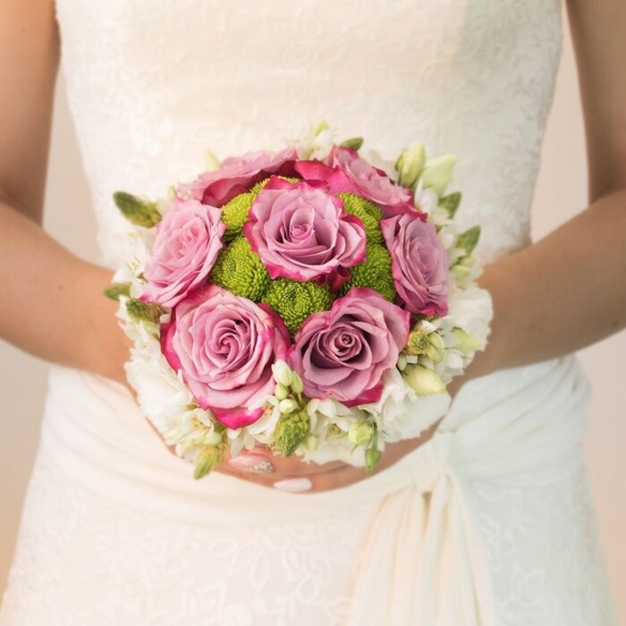 A timeless wedding moment unfolds in Gipsy Hill in this detailed close-up of a bride's bouquet, masterfully arranged in a compact, rounded shape that rests gently against a gown of intricate off-white lace. The central roses, enriched with layers of dusty mauve and purple-lavender, bloom fully-each petal delicately tipped in fuchsia, offering a play of colour that speaks to both romance and sophistication. Wrapped close to these are brilliant lime-green button chrysanthemums, their textured spheres introducing a fresh, playful element that echoes Gipsy Hill's parkland vibrancy. Framing the bouquet, a delicate scattering of petite white buds-freesia or spray roses-add a weightless, fragrant border. Subtle green buds and slender leaves further enliven the composition. The bride's hands, adorned with pale pink polish and a sparkling detail at the ring finger, cradle the bouquet with care. Filtered daylight gently highlights every fabric fold and floral cue, creating a scene infused with quiet elegance. As a creation suited for weddings across Gipsy Hill and Crystal Palace, this bouquet celebrates the harmony between traditional blooms and local personality.
