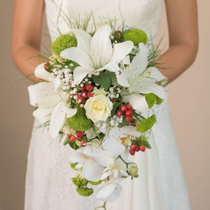 This intimate wedding scene from Gipsy Hill captures a close-up of a bride holding a lavish, cascading bouquet, crafted for modern romance with a nod to tradition. Majestic white lilies dominate the midsection, their trumpet blooms speckled delicately and evoking purity and new beginnings. A tightly swirled cream rose anchors the centre, surrounded by puffs of ice-white baby's breath that contribute a light, cloudlike aura. Sprays of vivid lime-green cushion chrysanthemums and clusters of gleaming red Hypericum berries bring fresh, festive flair, inspired by the seasonal palette visible in Gipsy Hill's community gardens and markets. Wispy, grass-like greenery and glossy dark foliage frame the bouquet, adding to its organic charm. Multiple white Phalaenopsis orchids trail downwards, creating the iconic teardrop cascade-a flourish reminiscent of water tumbling in Crystal Palace Park's fountains. The bride's gentle grip is visible against the delicate lace of her ivory gown, with the overall effect softened by a blurred, neutral background. Every flower and berry is rendered in sharp detail, celebrating the expertise of local florists and embodying the elegance that defines weddings in Gipsy Hill and Sydenham. This arrangement stands as an exquisite example of classic floral artistry heightened by modern touches and community spirit.